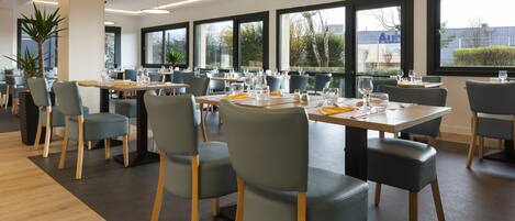 Un espace repas moderne doté de plusieurs tables et chaises, de grandes fenêtres et de plantes d’intérieur.