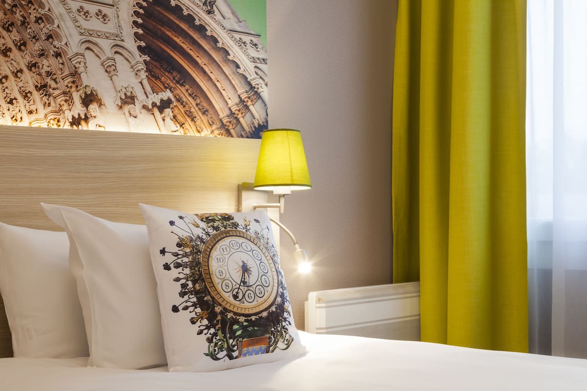 Habitación de hotel con cama, un cojín decorativo, un tapiz en la pared, una lámpara y cortinas.