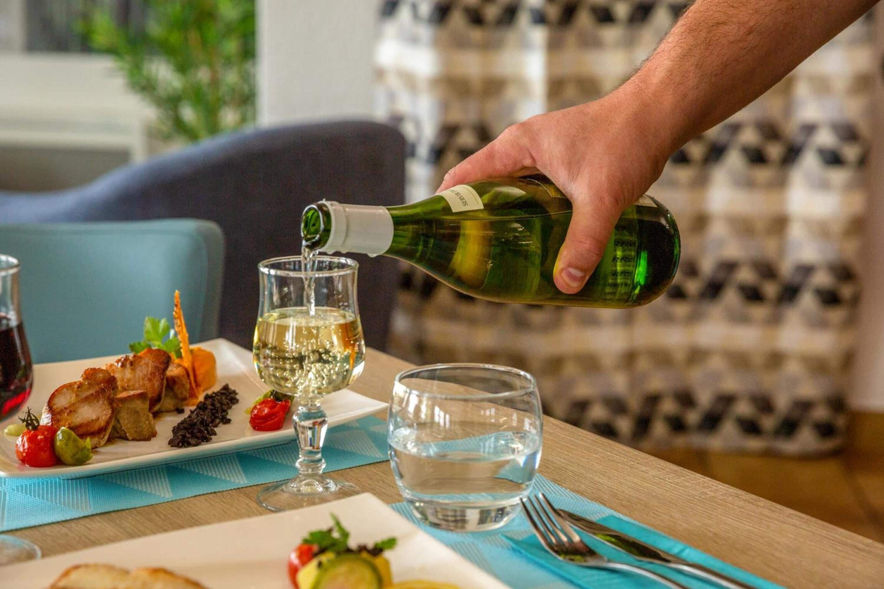 Una mesa de comedor con un plato de comida, una copa de vino blanco y una botella de vino siendo servido.