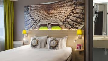 A hotel room with a bed, two bedside lamps, a wall mural, a nightstand, and a bathroom mirror.