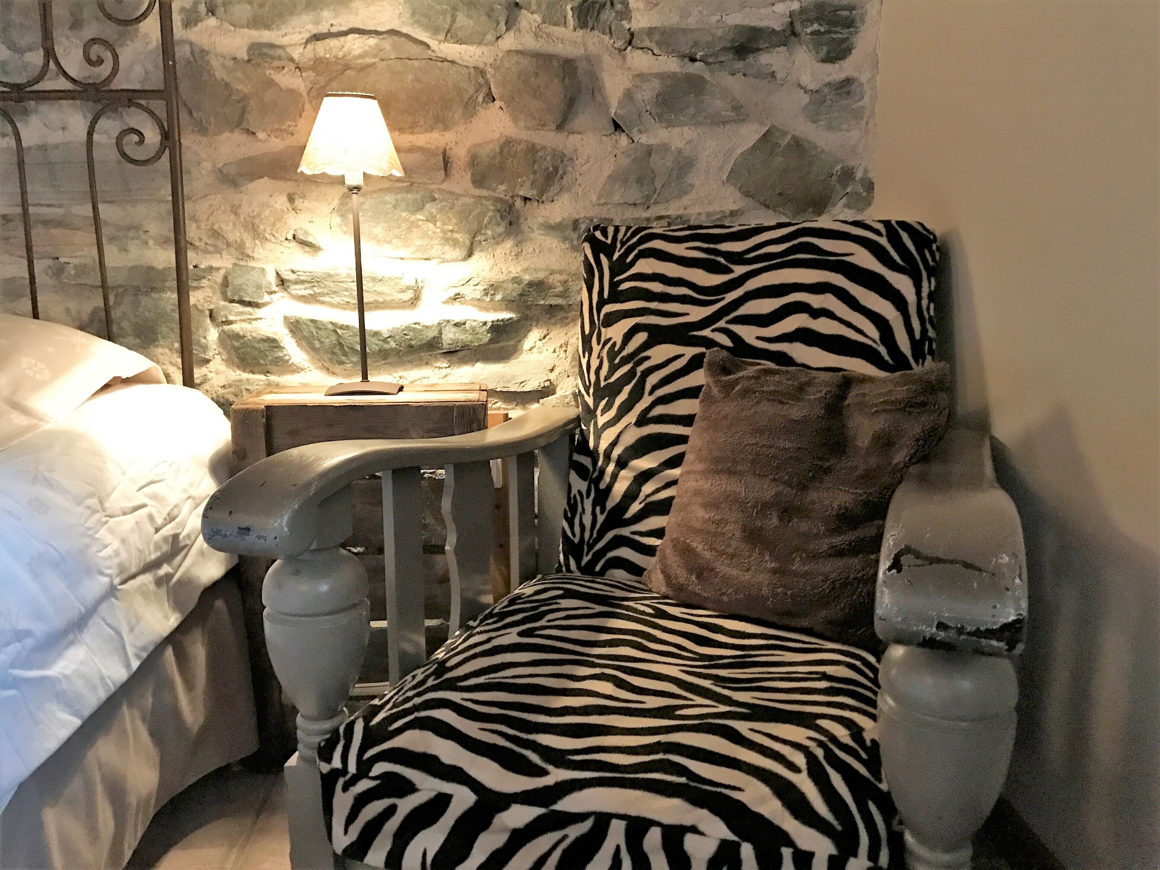 A bedroom with a stone wall, a bed with white bedding, a zebra-patterned chair, and a lamp on a wooden side table.