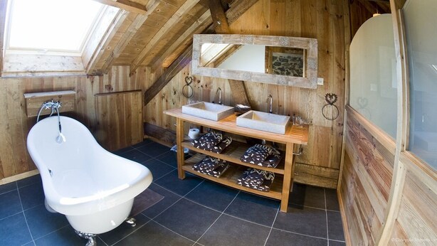Une salle de bain située dans les combles, avec un revêtement de sol en bois, une baignoire sur pieds, un meuble-lavabo en bois doté de deux vasques et un miroir au-dessus du meuble-lavabo.