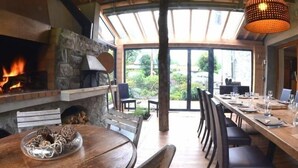 Un espace repas agrémenté d’une cheminée, de meubles en bois et offrant une vue à travers de grandes portes vitrées.