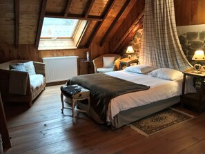 A cozy attic bedroom with a large bed, a wicker chair, a small stool, and a stone wall. - Chambres D'hôtes - La Clé des Bois (Le Bourg-d'Oisans)