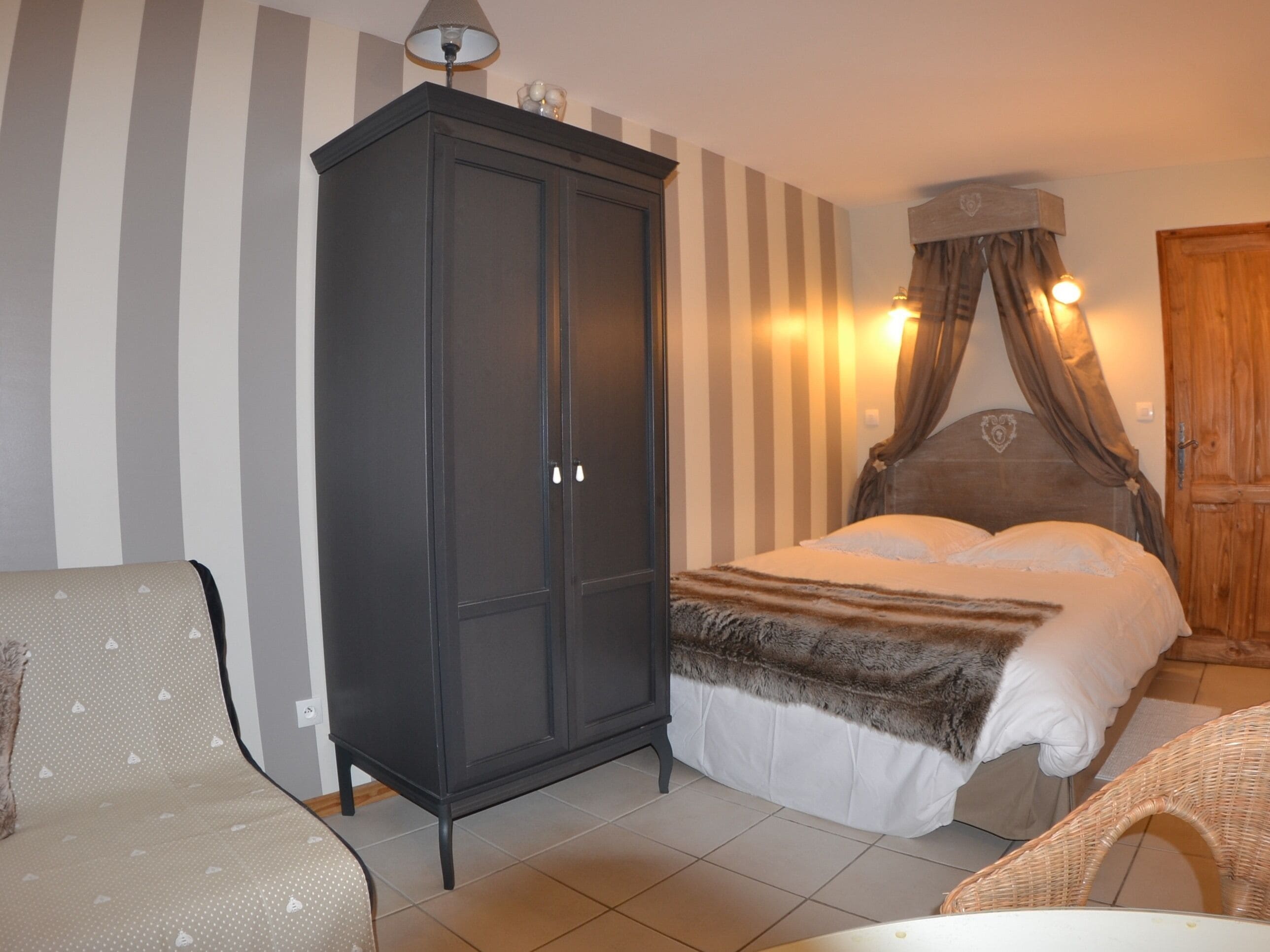 A bedroom with a large wardrobe, a bed with a canopy, a chair, and a wooden door.