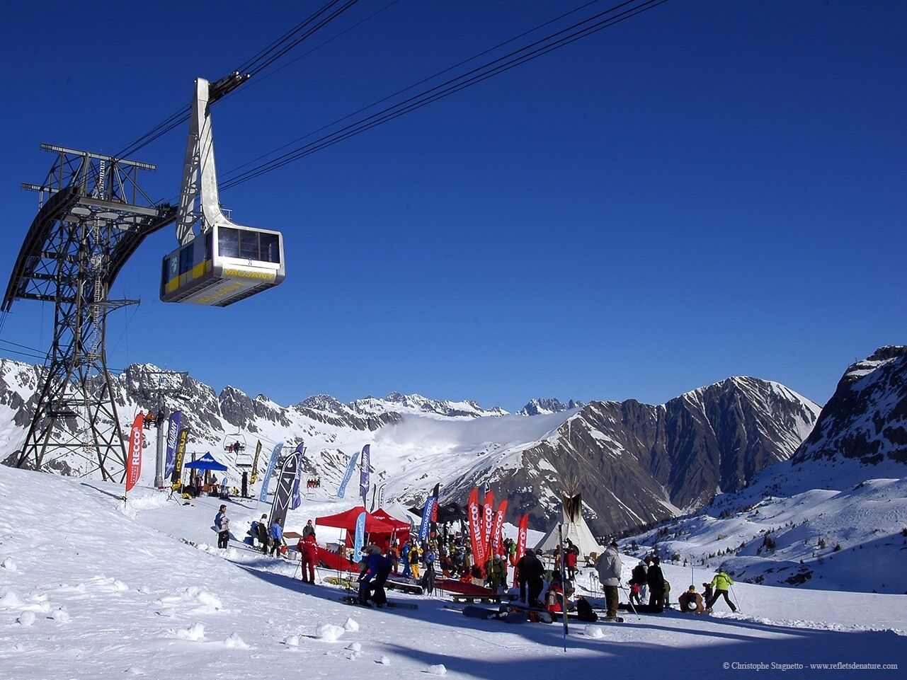 A ski lift with a gondola carrying passengers, a ski slope with people, and a mountainous backdrop.
