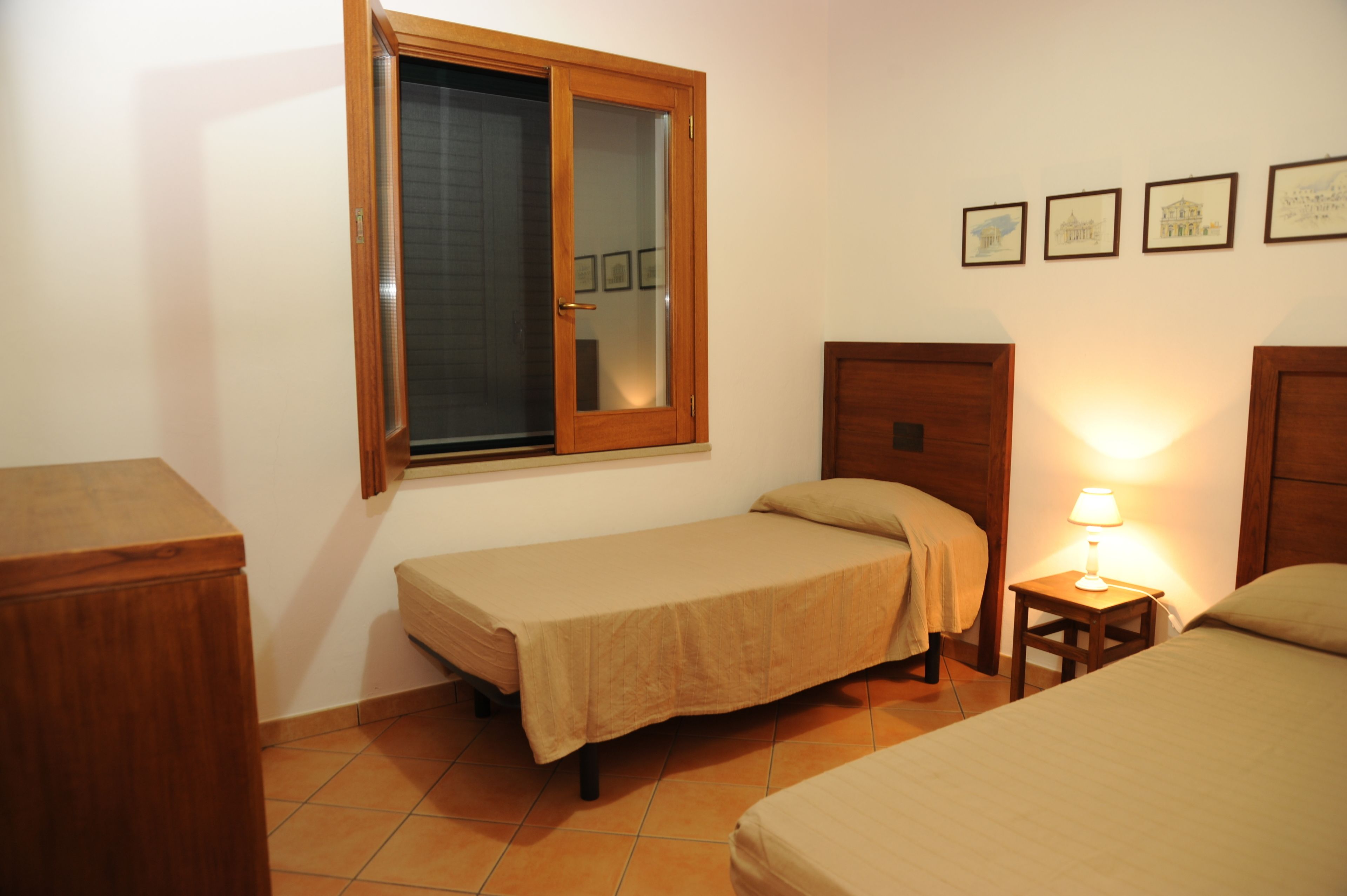 A small, tidy room with a single bed, a wooden cabinet, a nightstand with a lamp, and a window with a wooden frame.