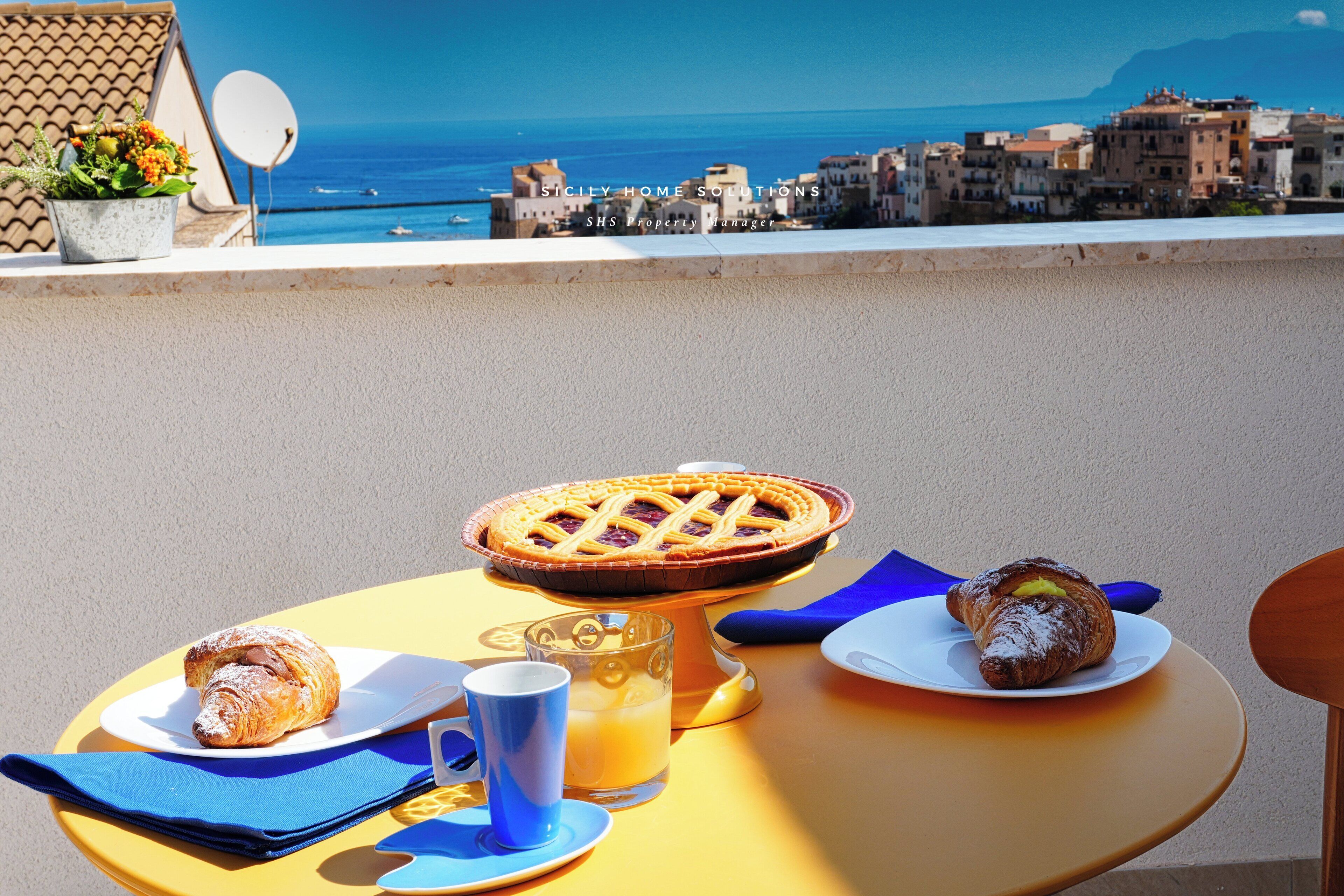 A table with a pie, a croissant, a mug, and a glass of orange juice, overlooking a coastal town.