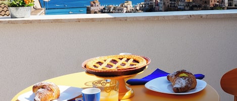 Mesa com uma torta, um croissant, uma caneca e um copo de suco de laranja, com vista para uma cidade costeira.