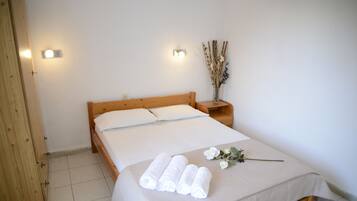 A neatly made bed with white linens, a wooden headboard, and a bedside table with a vase of dried plants.