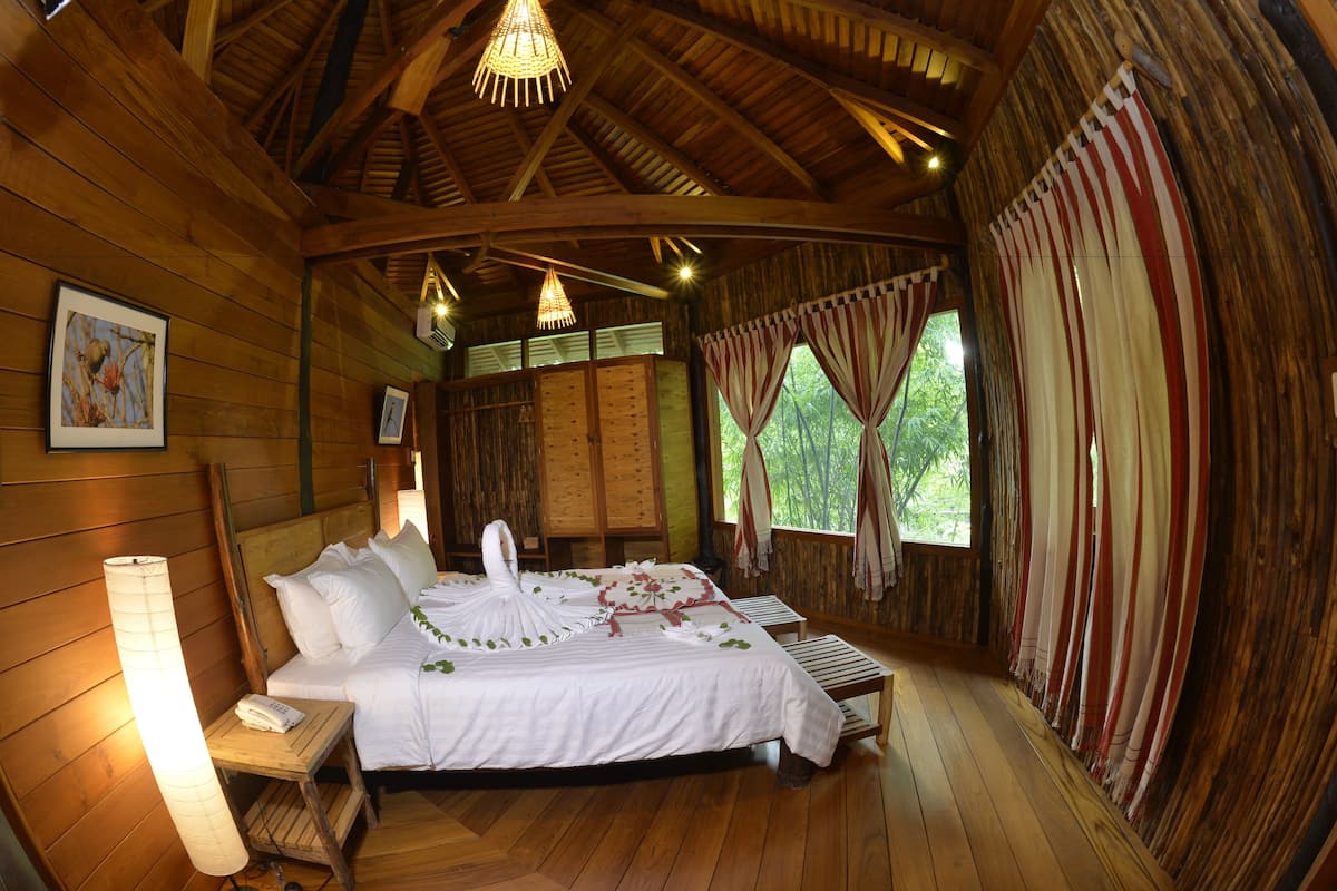 A wooden cabin room with a bed, a bedside lamp, and a window with curtains.