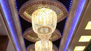 Un candelabro con diseños intrincados y varios niveles, suspendido de un techo con paneles decorados e iluminación azul.