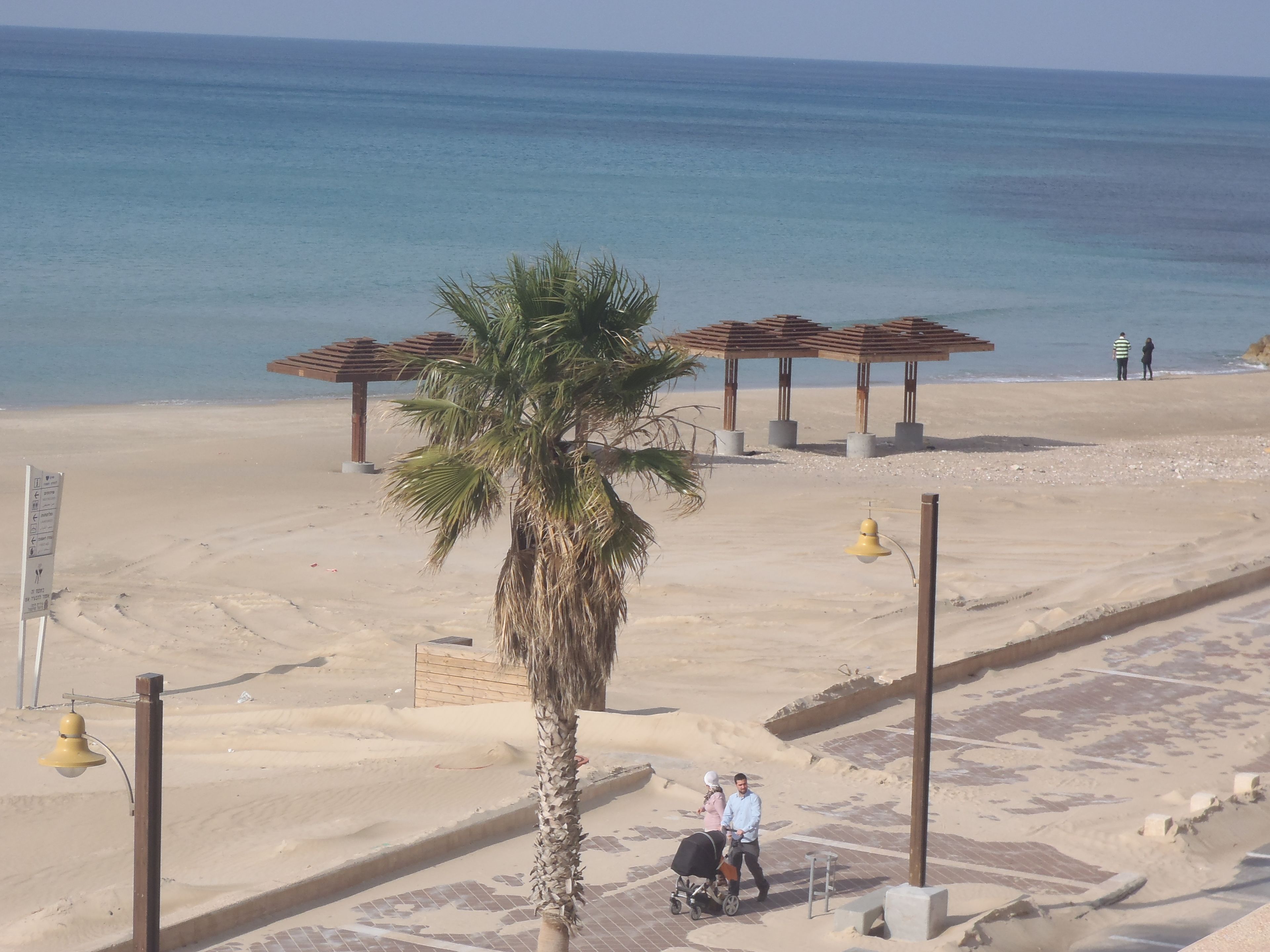 A beach with a palm tree, a wooden gazebo, and a couple with a stroller.