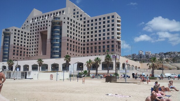 A large hotel with multiple floors and balconies, a sandy beach in the foreground, and palm trees. - Almog Studio (Haifa)