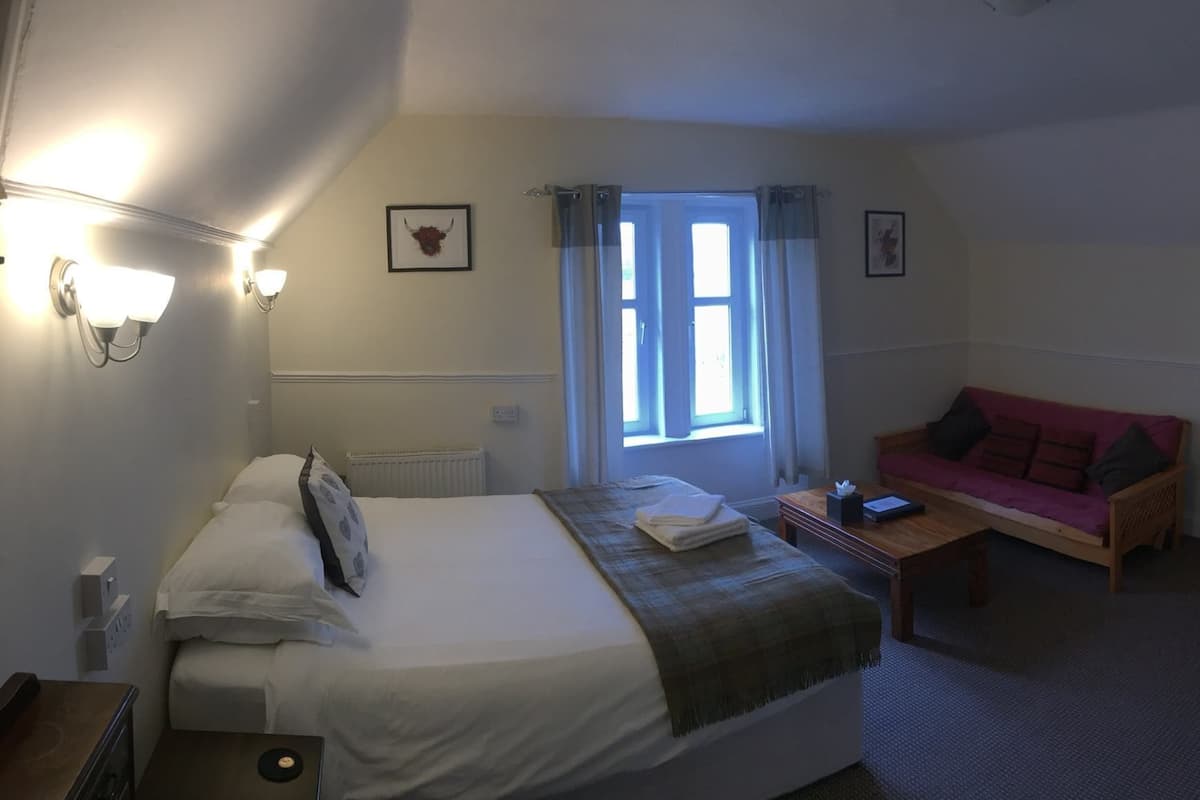 A bedroom with a bed, a bedside table, a window with curtains, a red sofa, and wall-mounted lights.