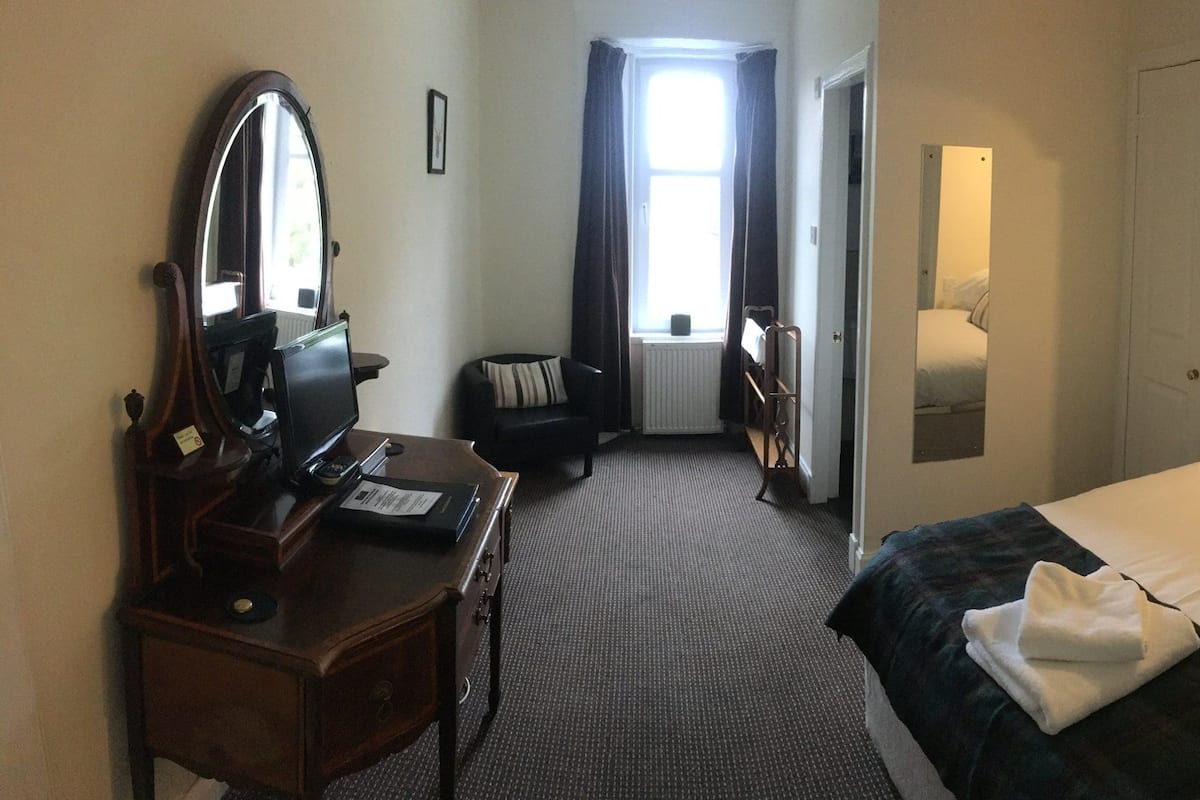A hotel room with a bed, a desk with a mirror, a chair, and a window with curtains.
