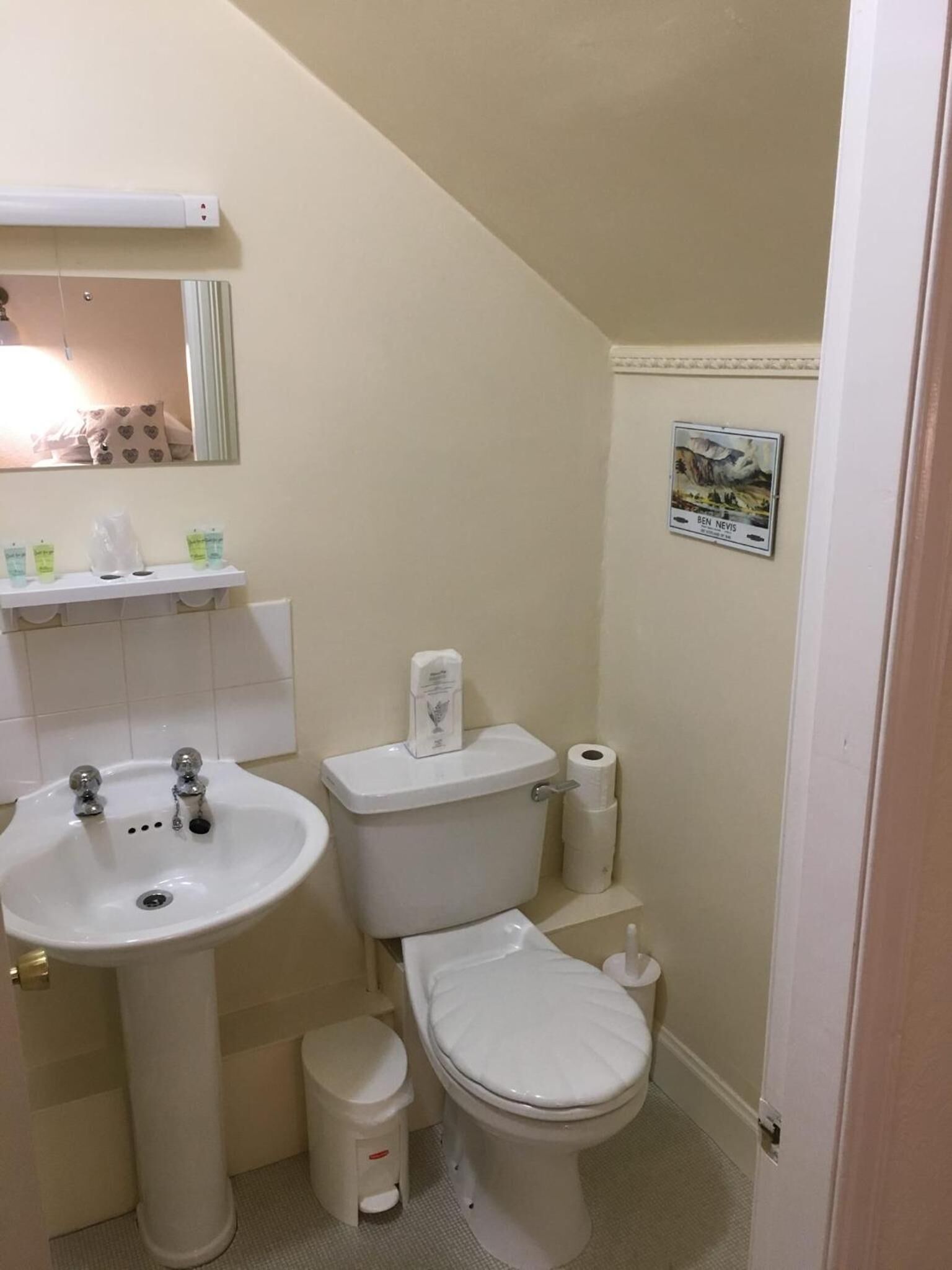 A small bathroom with a white sink, toilet, and bidet. There is a mirror above the sink and a small shelf with toiletries.