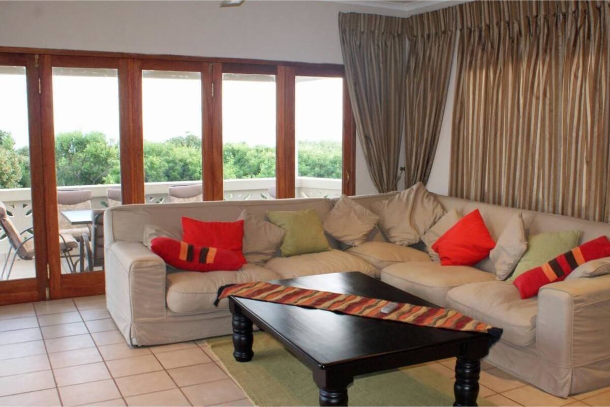A living room with a beige sofa, red and green cushions, a black coffee table, and large windows with curtains.