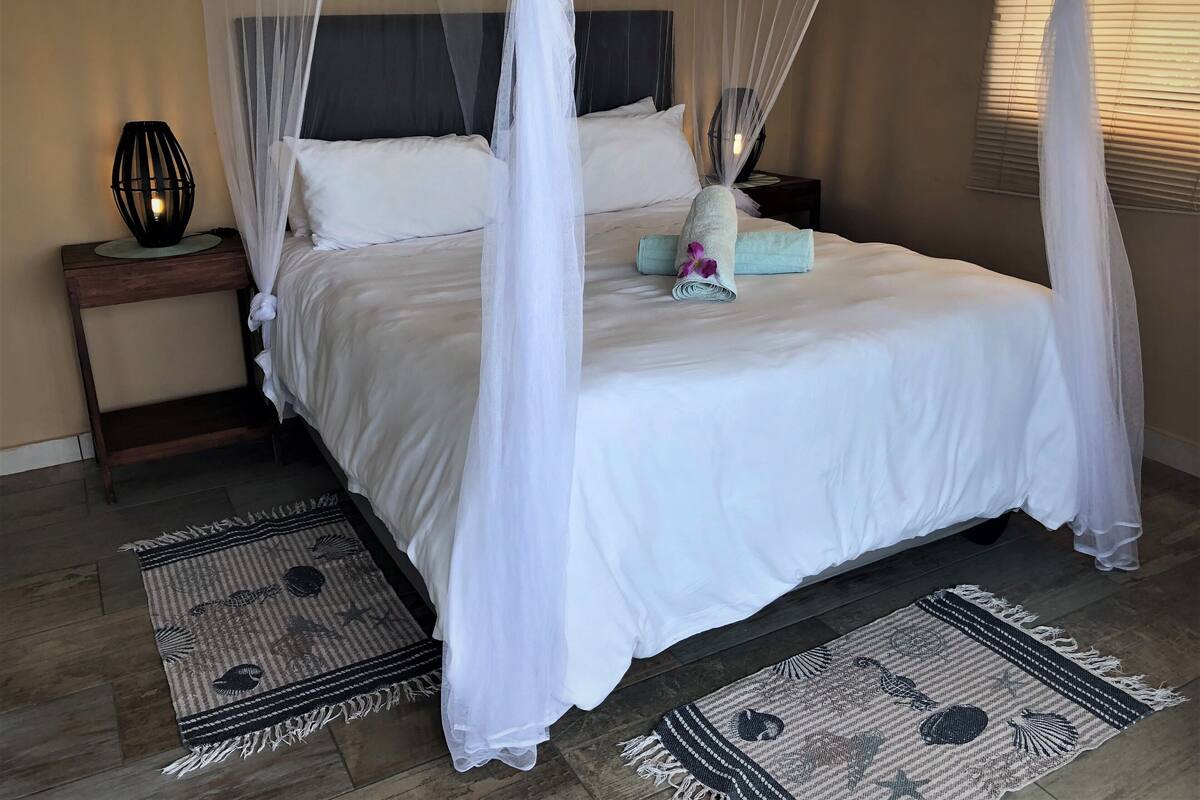A neatly made bed with white linens, a decorative pillow, and a rolled towel. There is a nightstand with a lamp and a small vase with a flower. The room has a wooden floor and two patterned rugs.