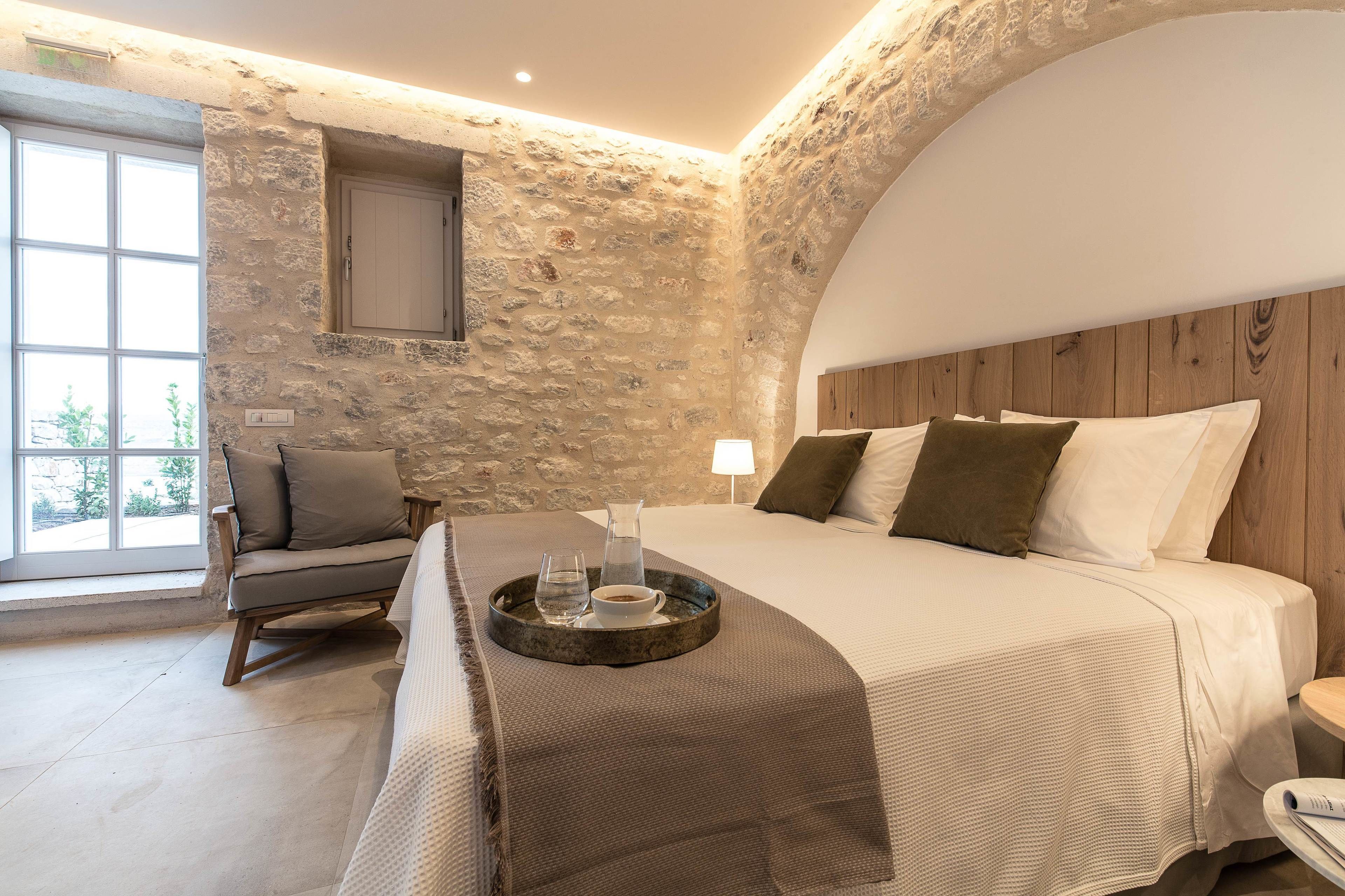 A bedroom with a stone wall, a wooden headboard, a bed with white bedding, a side table, a chair, and a window with a view.