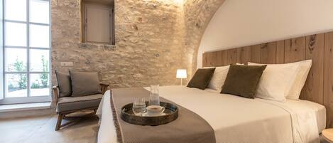 A bedroom with a stone wall, a wooden headboard, a bed with white bedding, a side table, a chair, and a window with a view.