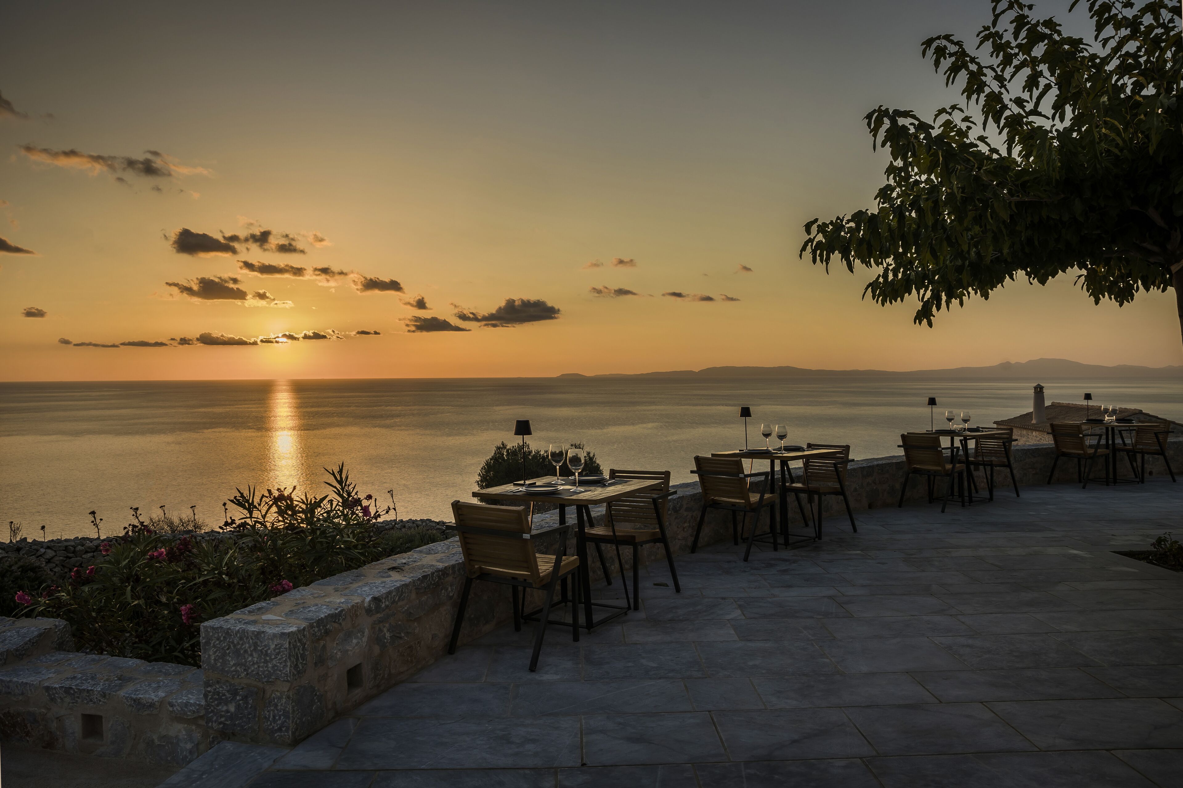 An outdoor dining area with tables and chairs overlooking a body of water during sunset.
