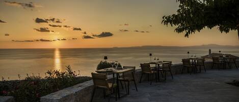 An outdoor dining area with tables and chairs overlooking a body of water during sunset.