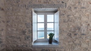 A stone wall with a window overlooking the sea, a potted cactus, and a cushioned bench. - Ariá Estate Suites & Spa (East Mani)