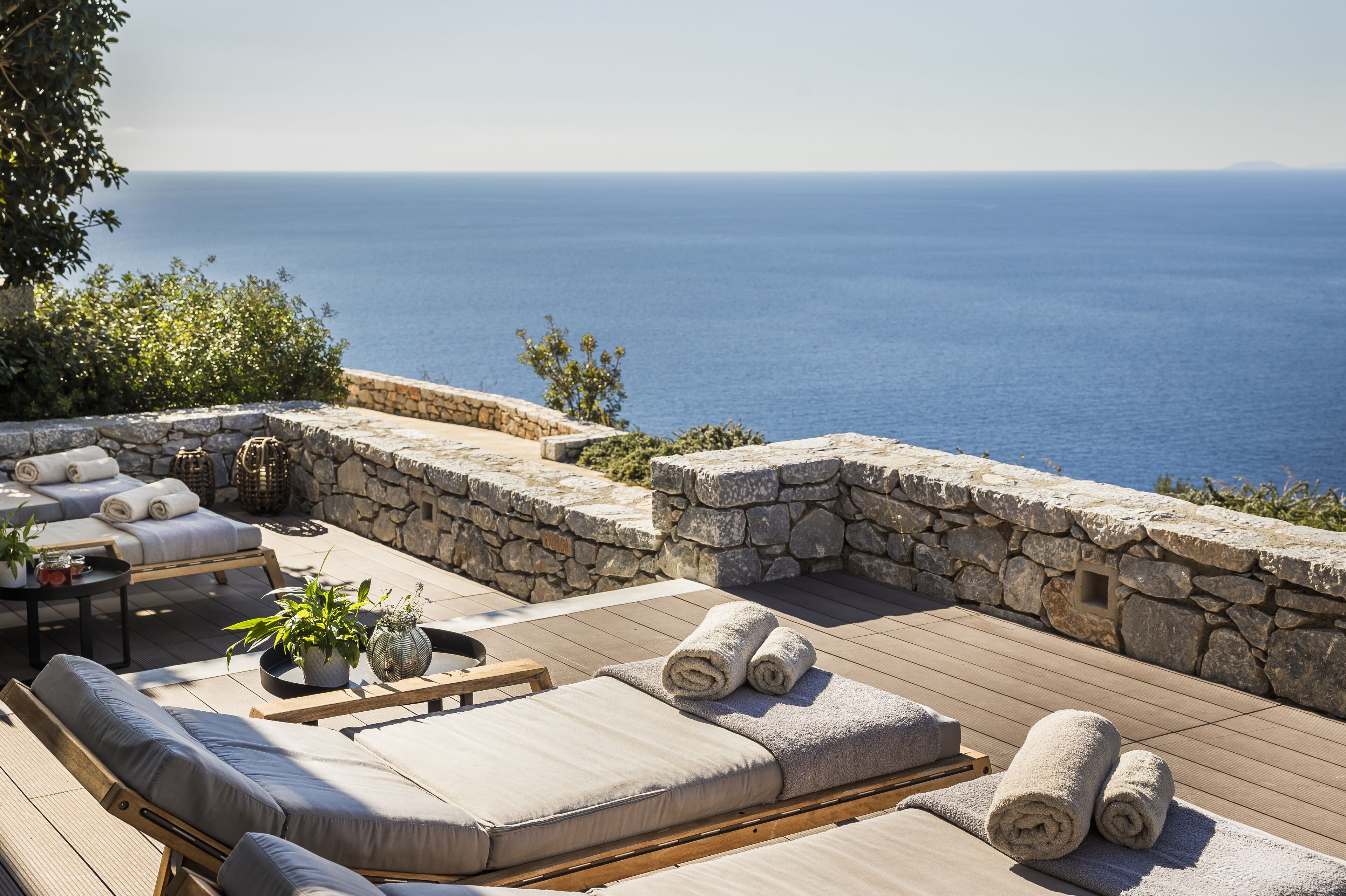 Un espacio de estar al aire libre con luz solar, paredes de piedra, una terraza de madera y vistas al mar.