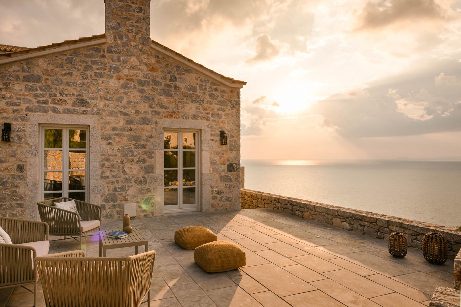 A stone building with a terrace overlooking the sea, featuring wicker furniture and a view of the sunset.