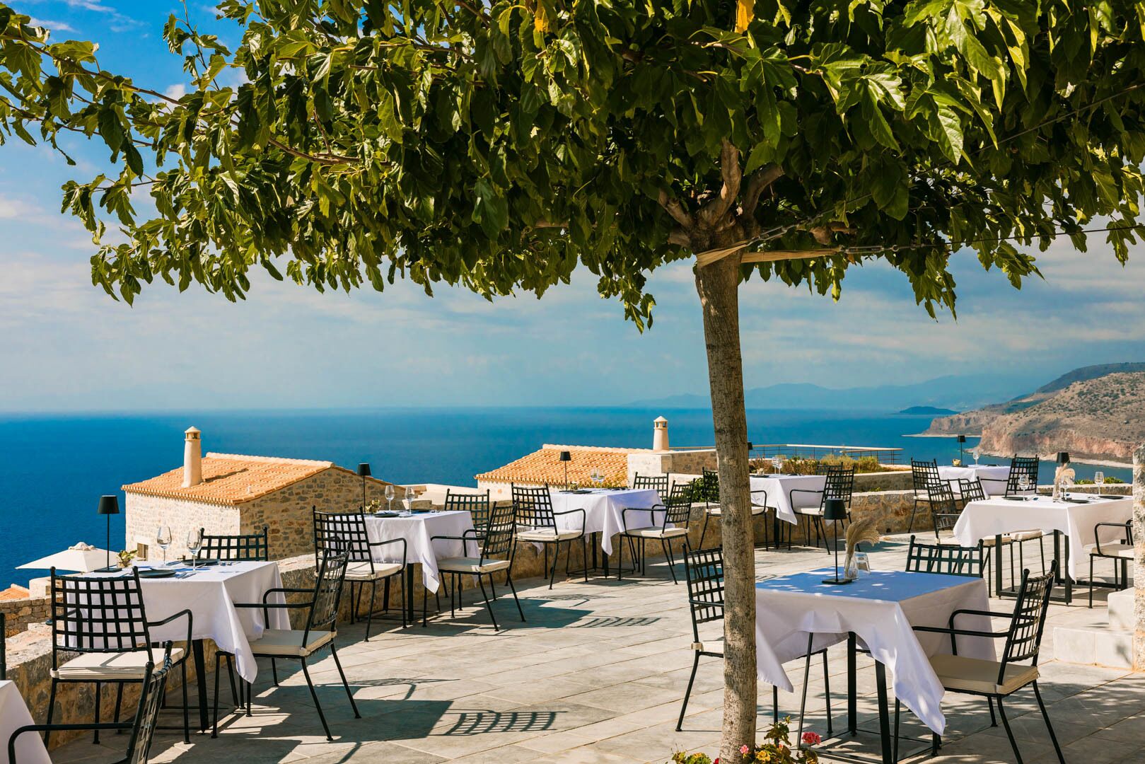 An outdoor dining area with tables and chairs, overlooking a scenic view of the sea and coastline.
