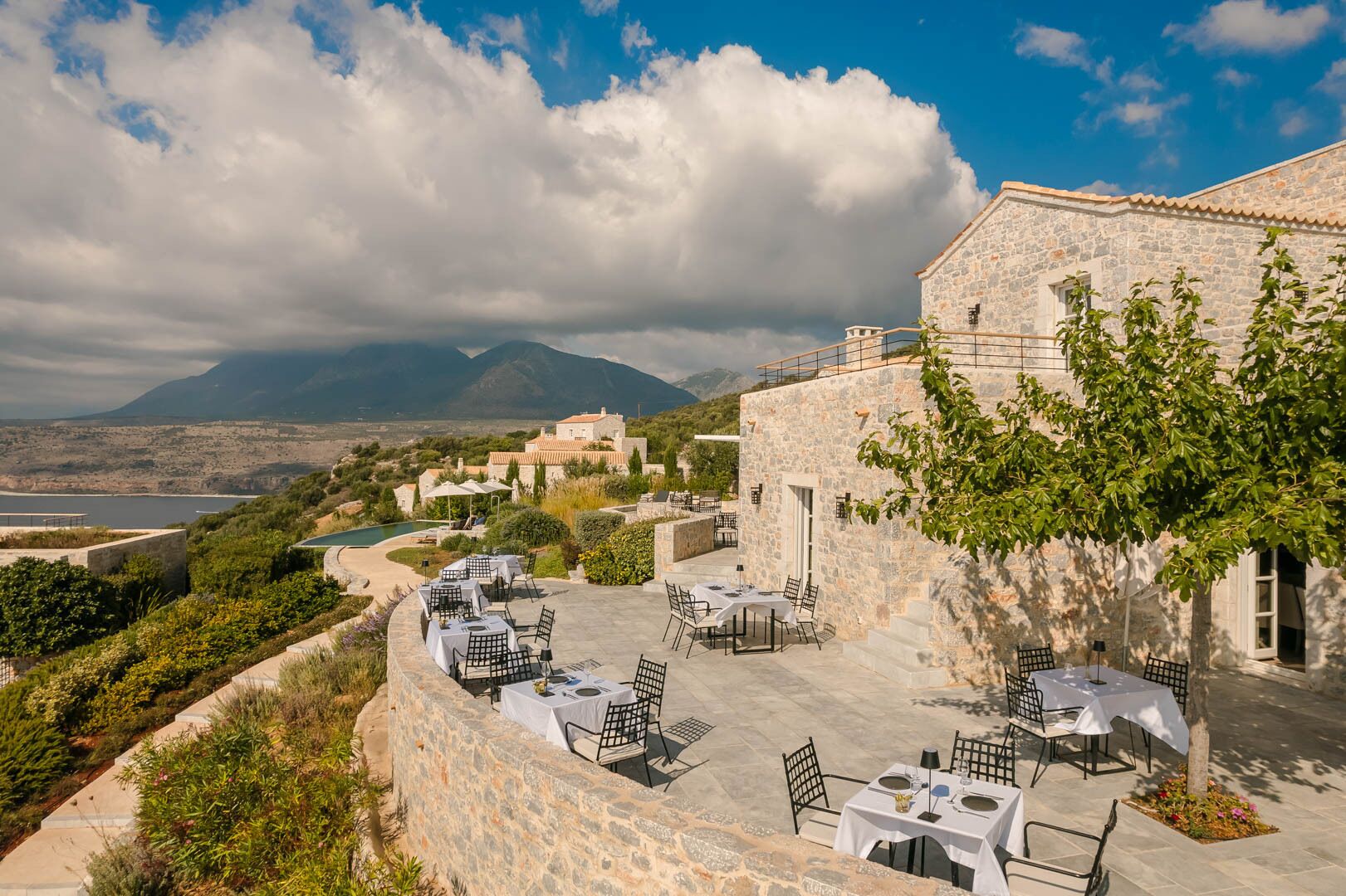 Un área de comedor al aire libre con paredes de piedra y mesas, con vistas a un paisaje montañoso y un cuerpo de agua.