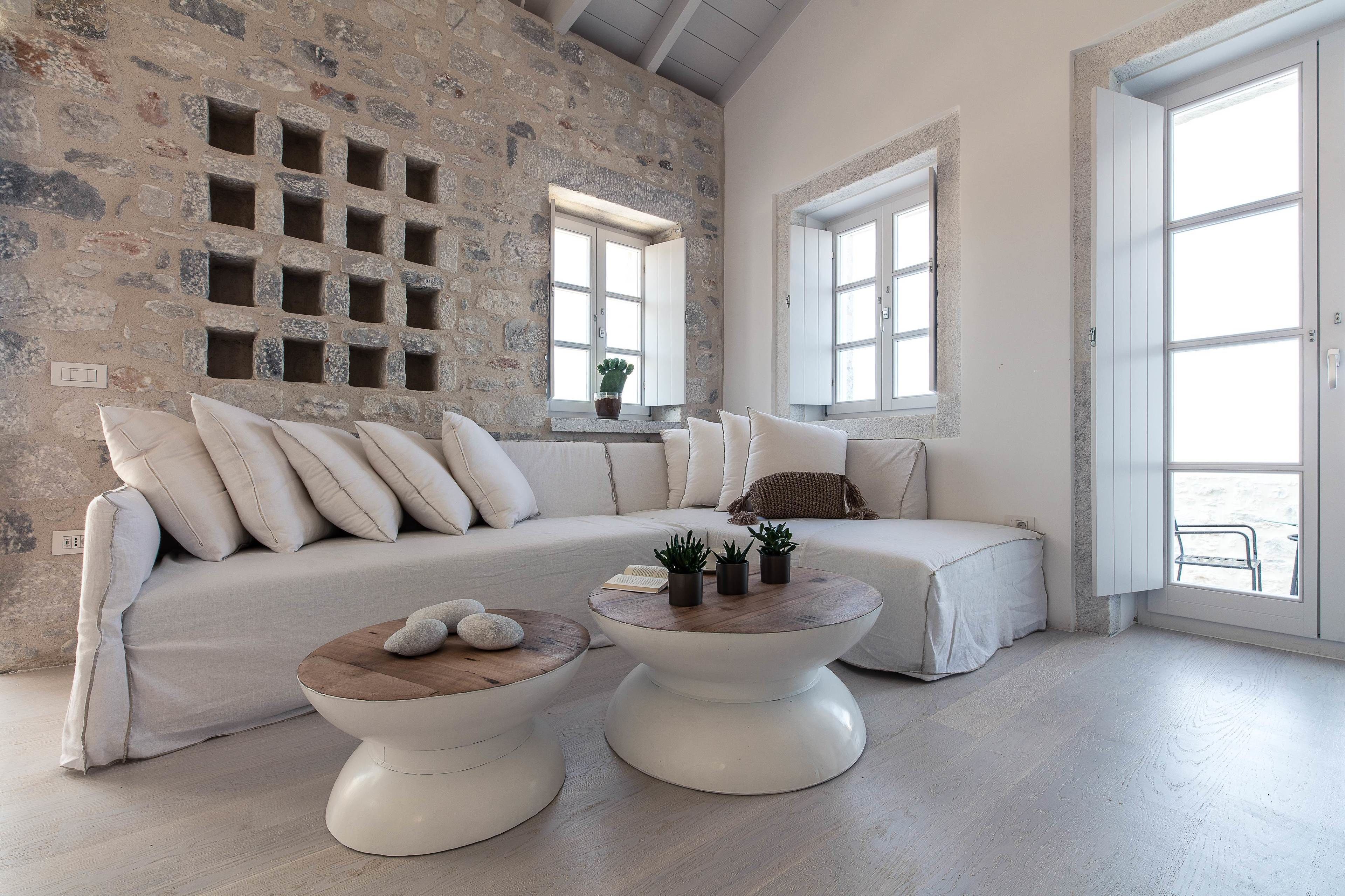 A modern living room with a stone wall, a white sofa, two white coffee tables, and a large window.