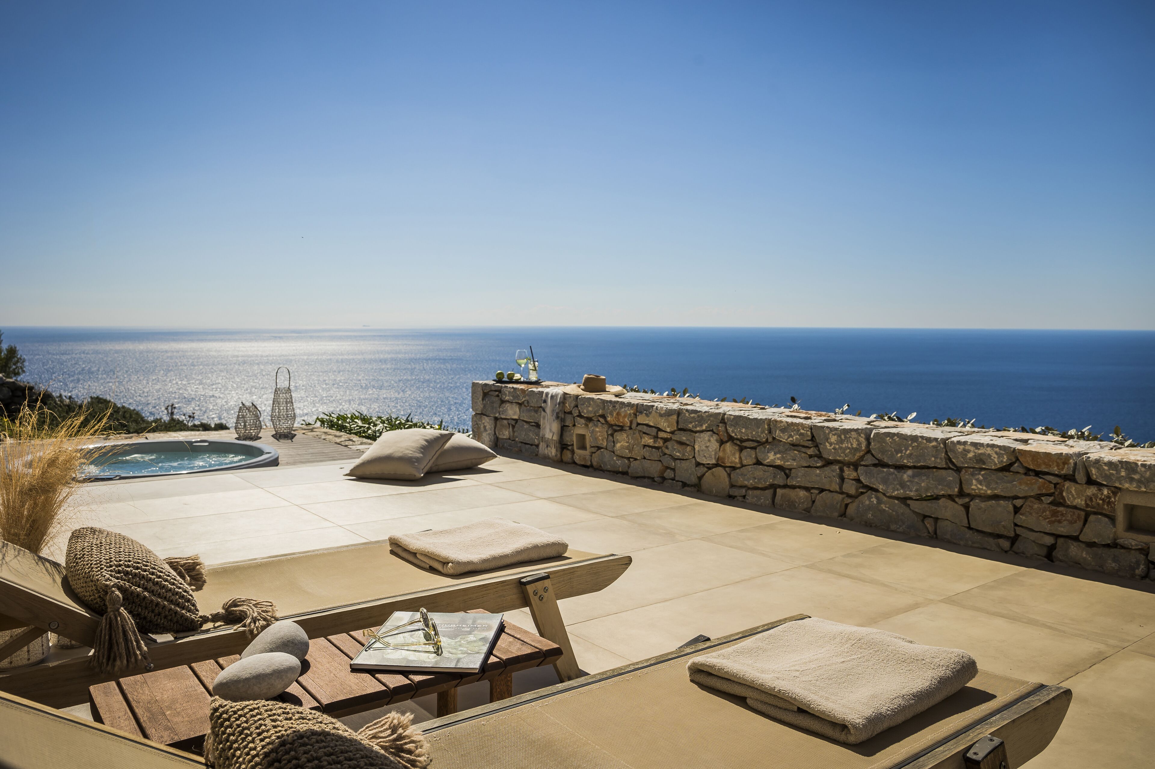 A sunlit outdoor lounge area with stone walls, a hot tub, and lounge chairs overlooking the sea.
