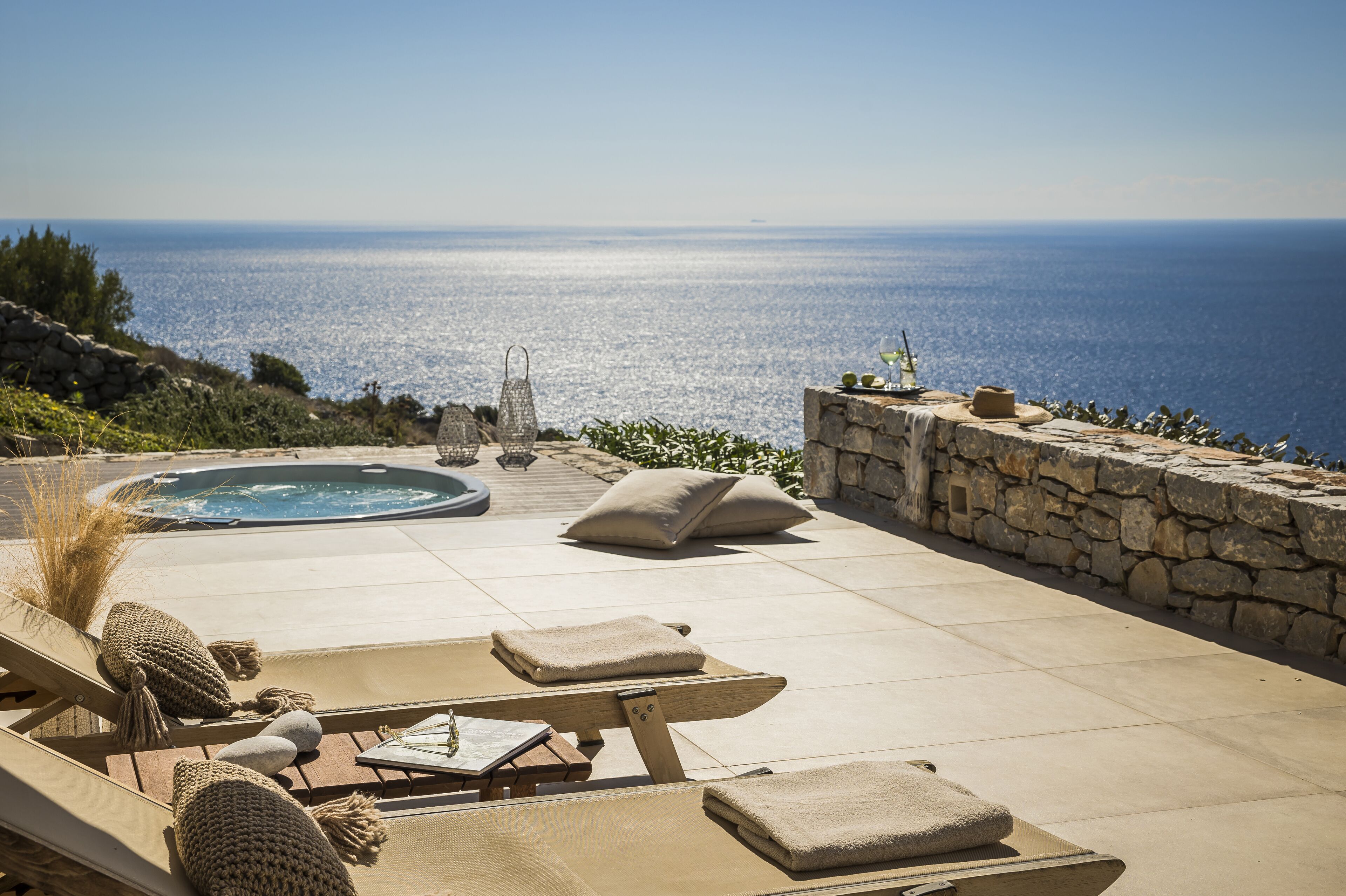 A sunlit outdoor seating area with lounge chairs, a hot tub, and a stone wall overlooking the sea.