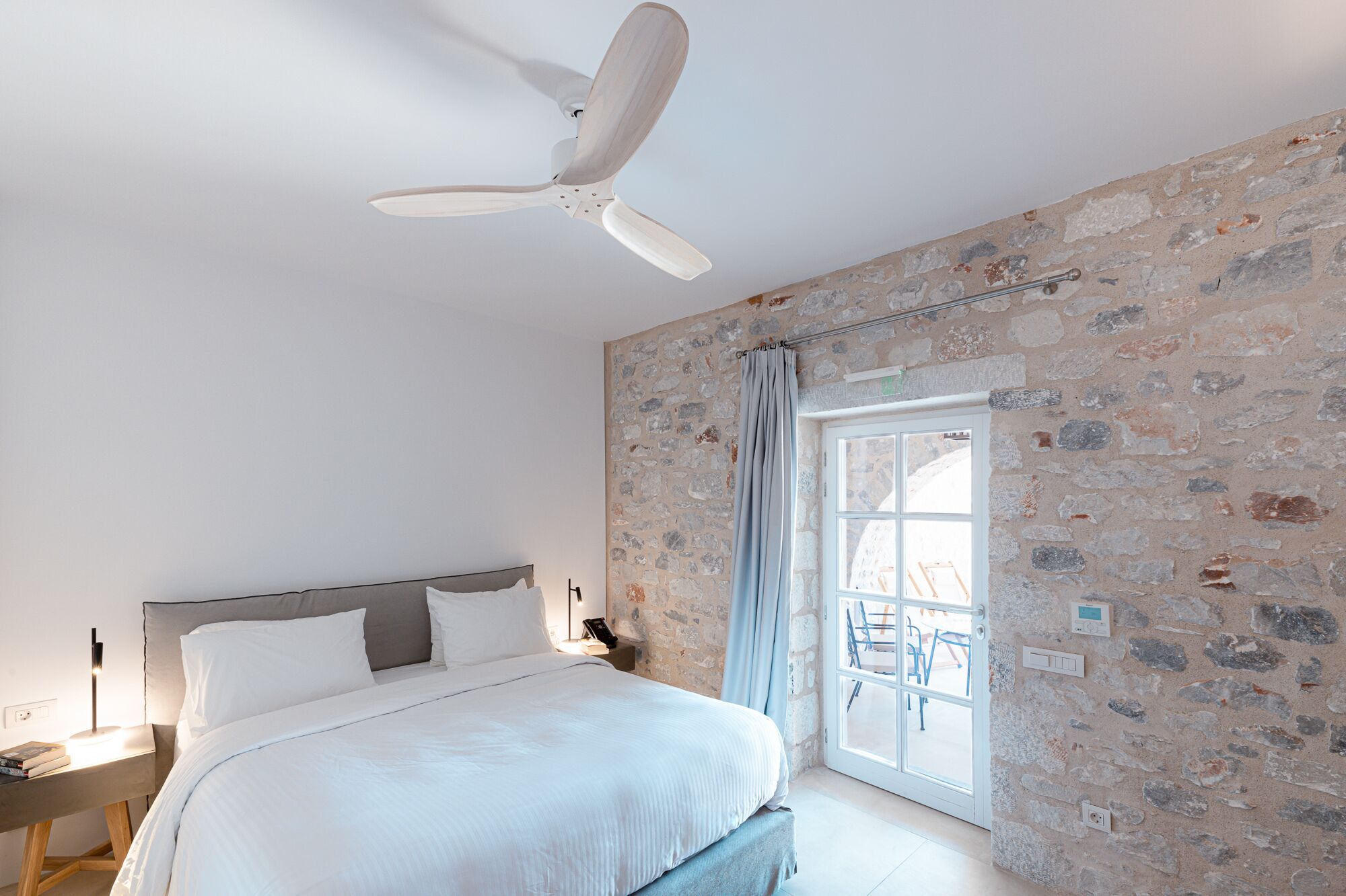 A bedroom with a bed, a ceiling fan, a stone wall, and a window with a view.
