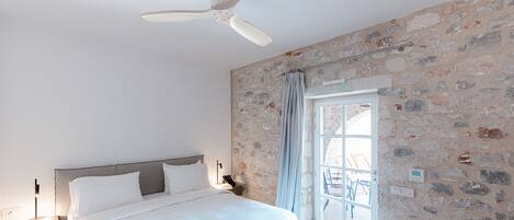 A bedroom with a bed, a ceiling fan, a stone wall, and a window with a view.