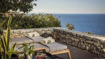 Relax in a serene outdoor space overlooking the sea, surrounded by lush greenery and a rustic stone wall.