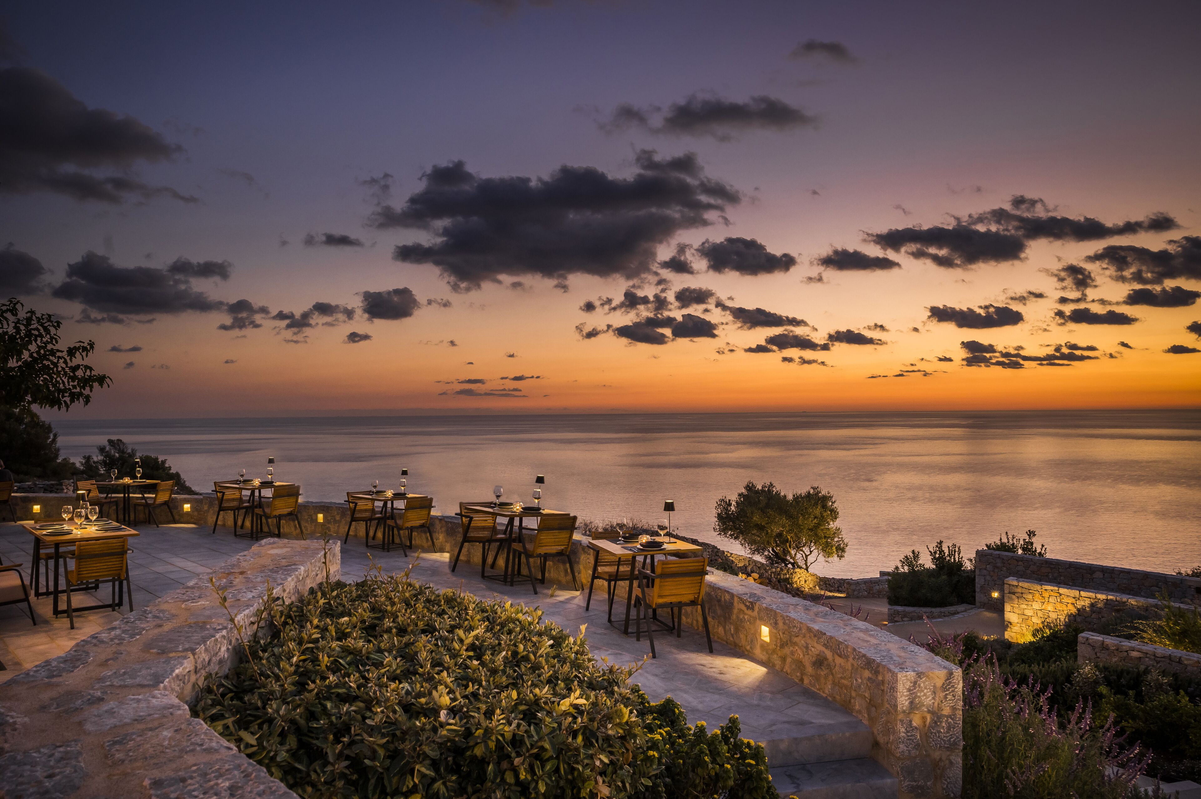 An outdoor dining area with tables and chairs, overlooking a body of water at sunset.