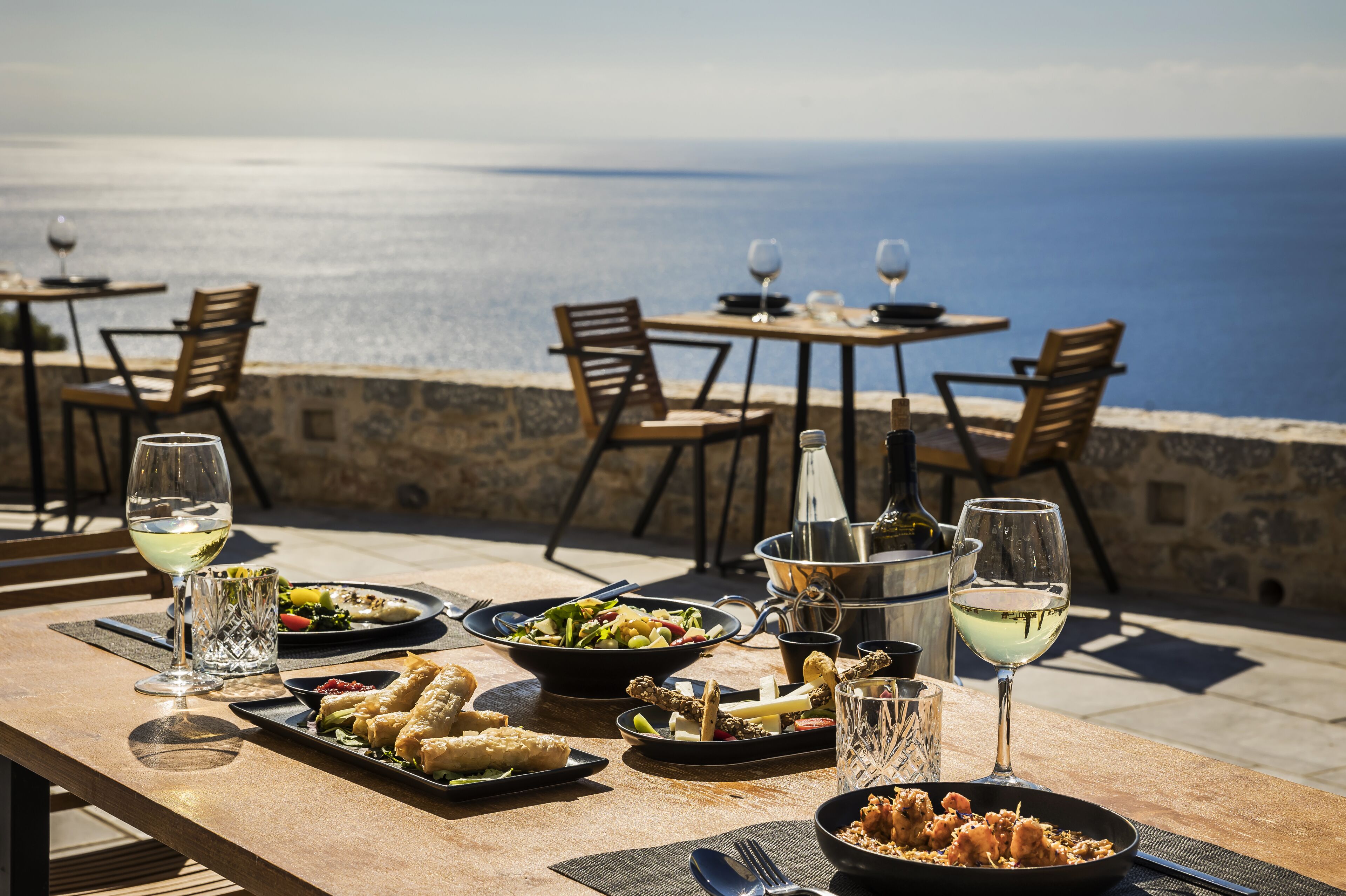 Un espace de restauration en plein air, avec des tables et des chaises, une variété de plats et des verres à vin, offrant une vue sur une étendue d’eau.