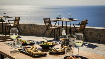 Un espace de restauration en plein air, avec des tables et des chaises, une variété de plats et des verres à vin, offrant une vue sur une étendue d’eau.