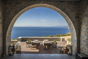 A stone archway overlooking a wooden deck with two lounge chairs and a table, set against a backdrop of the sea and distant mountains. - Ariá Estate Suites & Spa (East Mani)