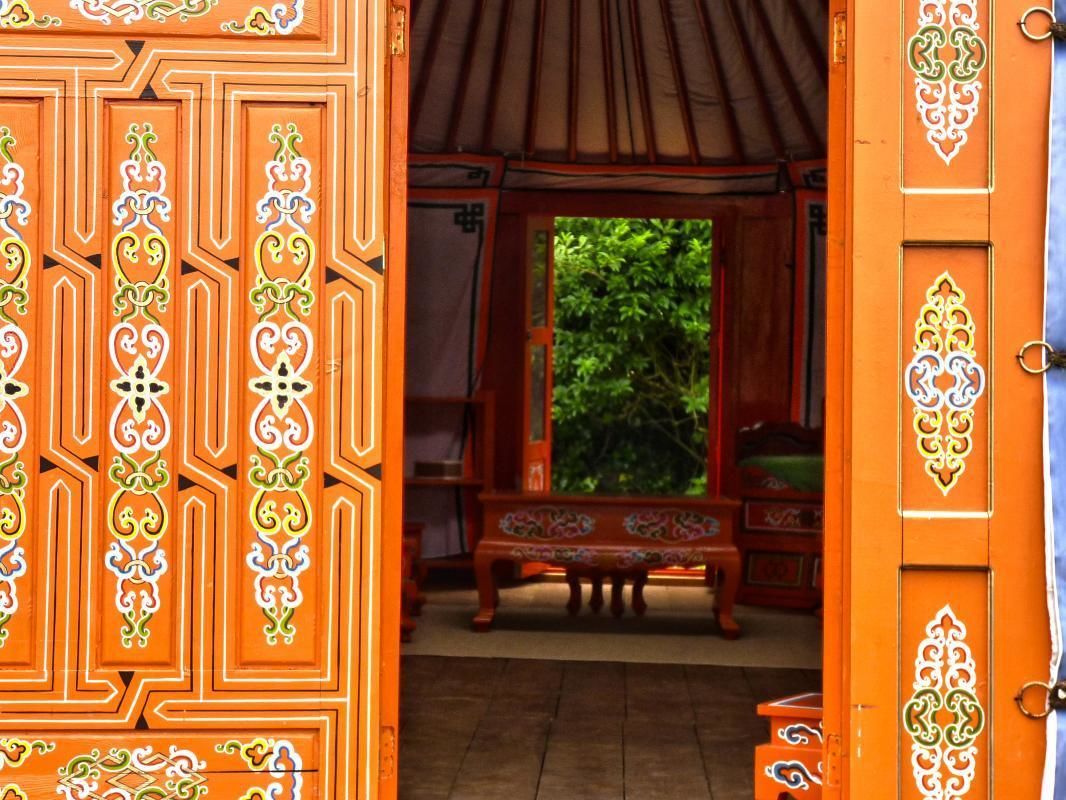 Una puerta de madera abierta con tallas intrincadas, que da a un cuarto con un banco y vistas al exterior con vegetación.