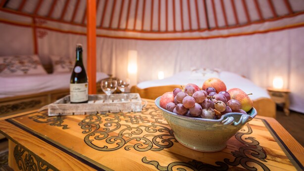Una mesa de madera con un bol de fruta y una botella de vino en una habitación tipo tienda, con una cama y una mesita de noche al fondo.