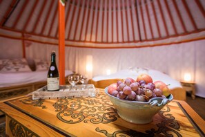 Una mesa de madera con un bol de fruta y una botella de vino en una habitación tipo tienda, con una cama y una mesita de noche al fondo.