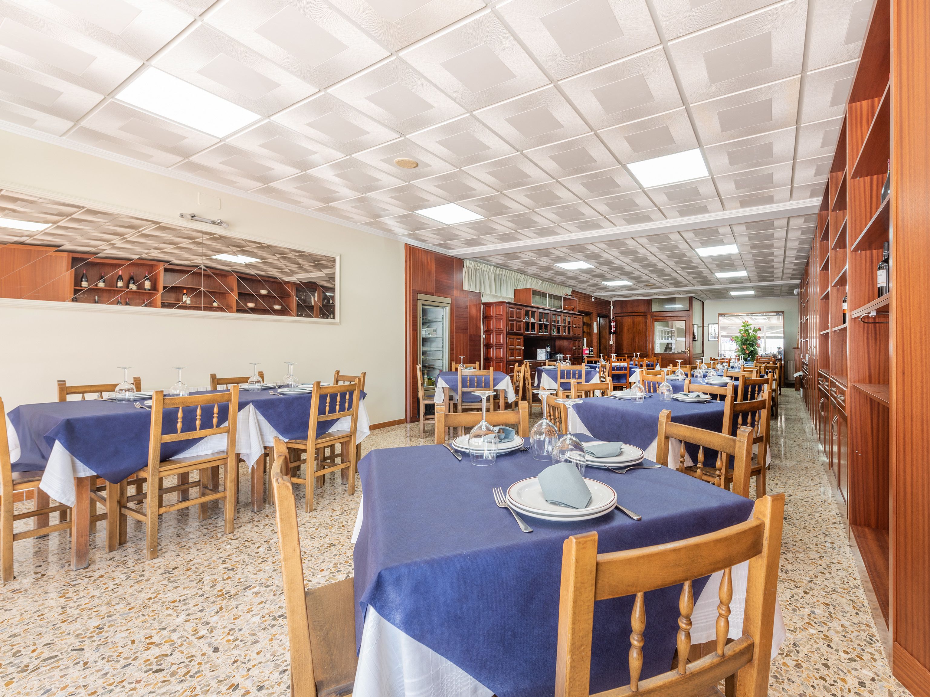A restaurant with multiple tables, each covered with a blue tablecloth and set with white plates and silverware. The ceiling has a grid pattern, and there are wooden cabinets along the walls.