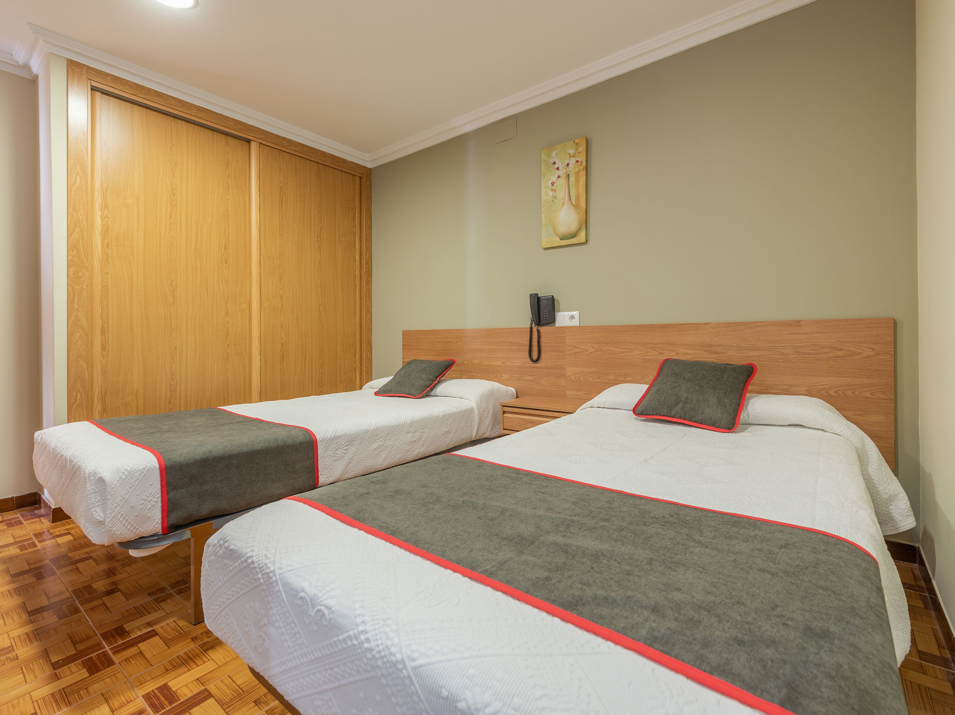 A hotel room with two beds, a wooden wardrobe, a painting on the wall, and a phone on the nightstand.