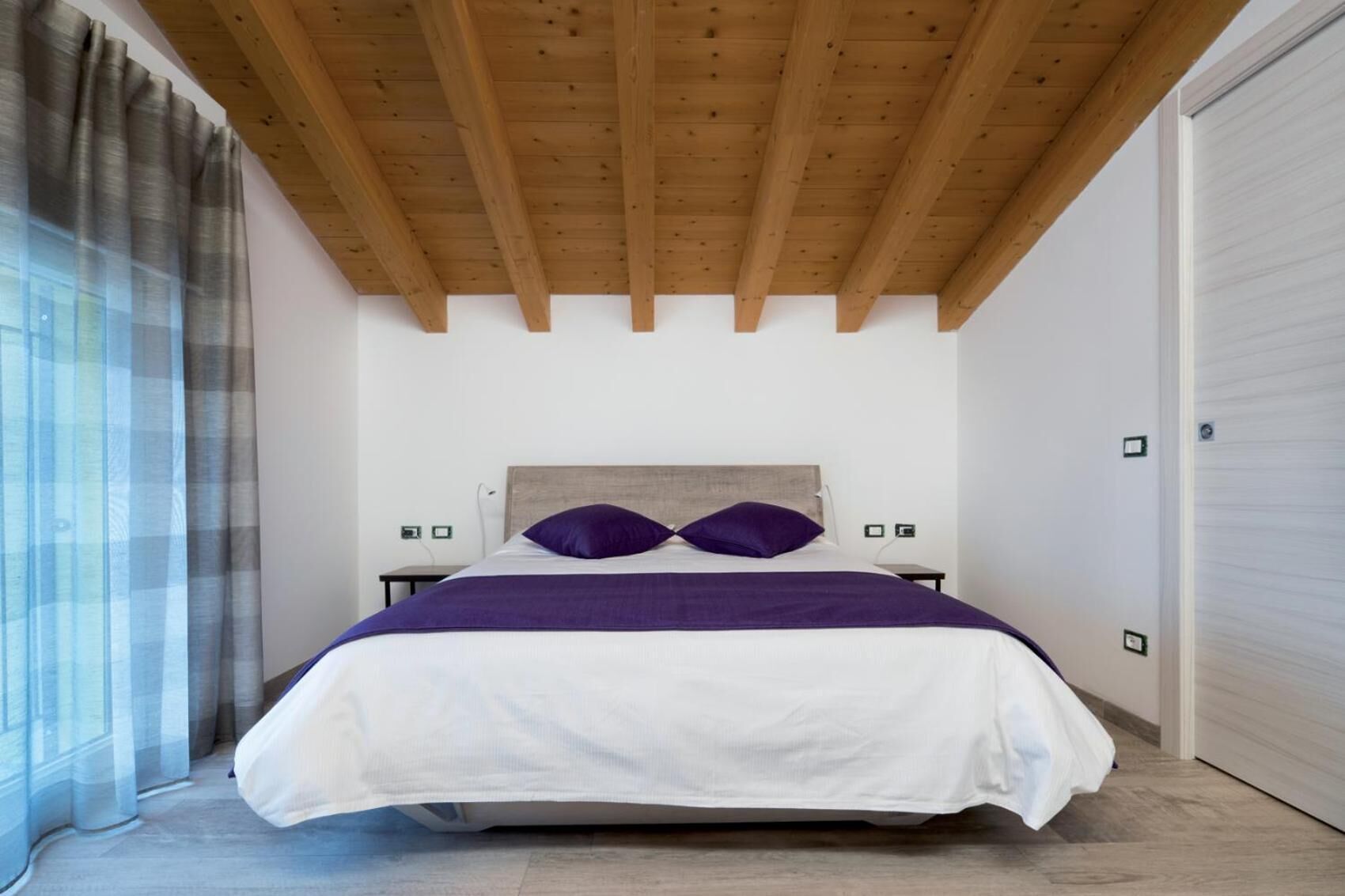 Un dormitorio con techo de madera, una cama con ropa de cama blanca y morada, una mesita de noche y una ventana con cortinas.
