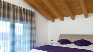 A bedroom with a wooden ceiling, a bed with white and purple bedding, and a window with sheer curtains. - Agriturismo Camisi (Cambiago)
