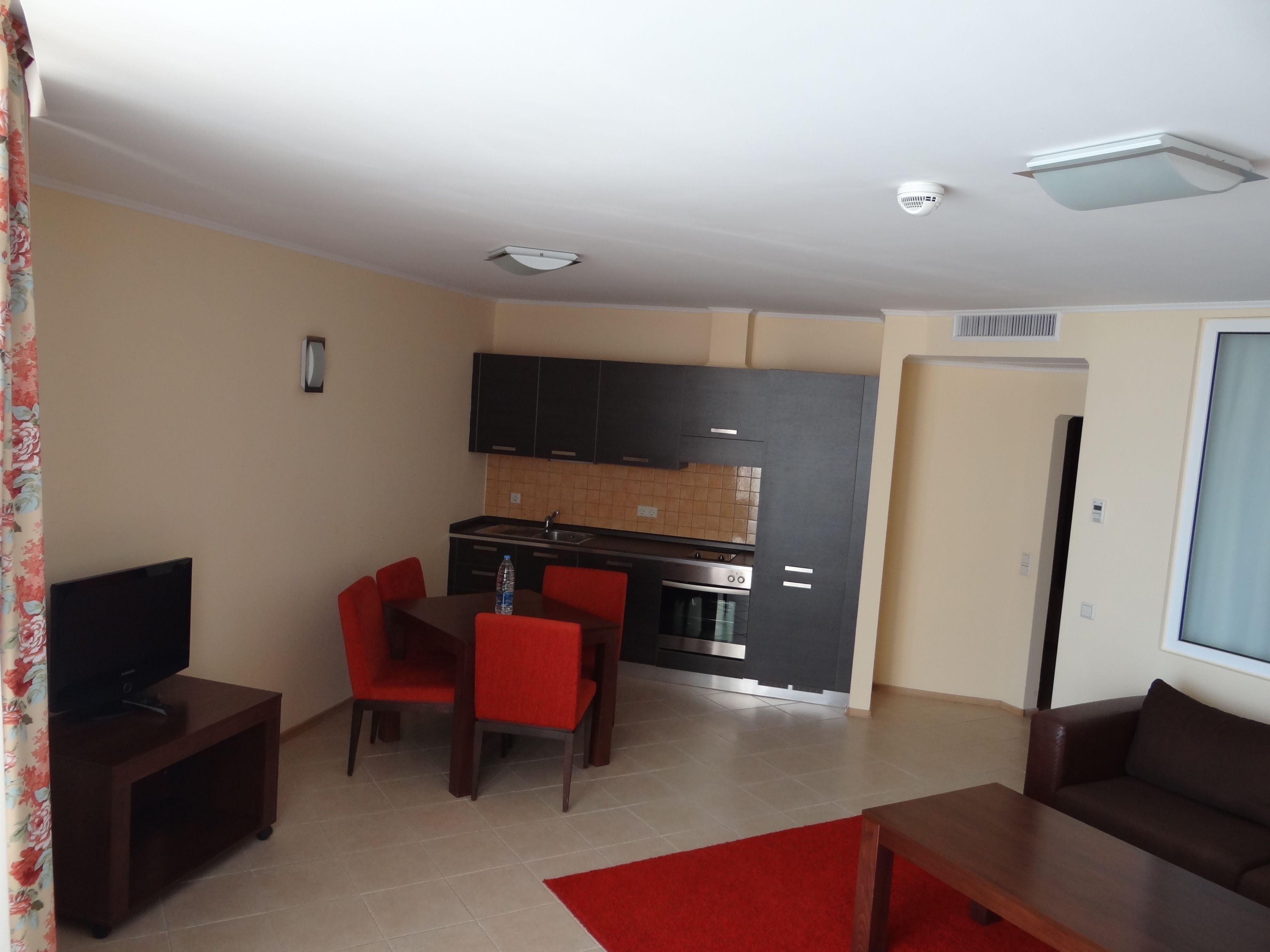 A compact living space with a kitchenette, a dining area with red chairs, a TV on a stand, and a sofa.